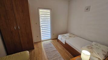 Quarto pequeno e bem iluminado, com guarda-roupa de madeira, uma cama de solteiro com colcha estampada, mesa de cabeceira com abajur e um tapete pequeno no chão de madeira.