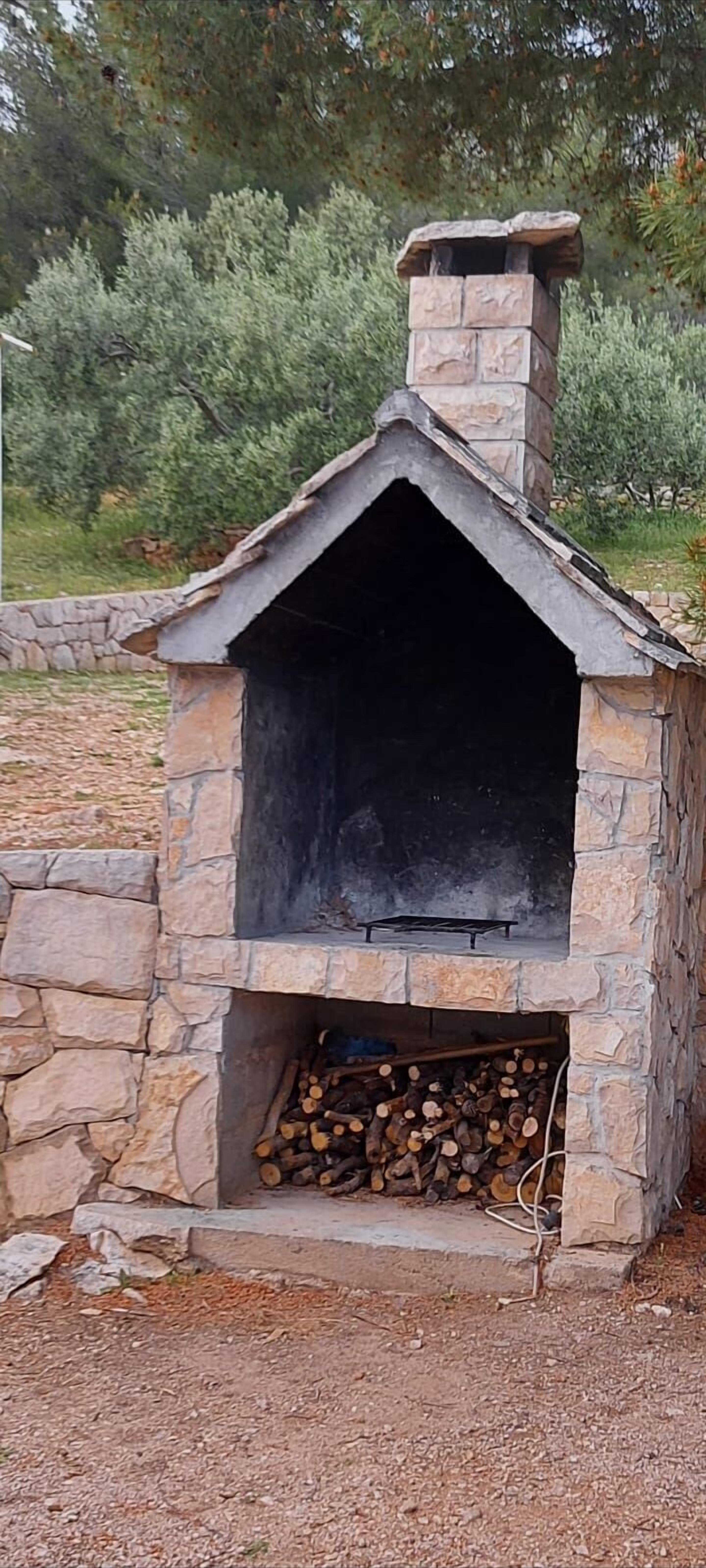 A stone-built outdoor fireplace with a chimney, surrounded by a stone wall and a pile of firewood.
