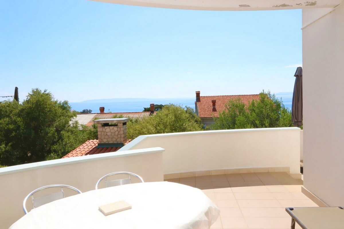 A balcony with a table and chairs, a view of the sea, and a stone structure in the background.