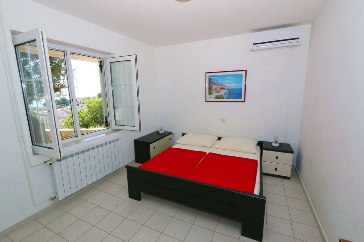 A bedroom with a bed, a window, a radiator, a nightstand, and a wall-mounted air conditioner.