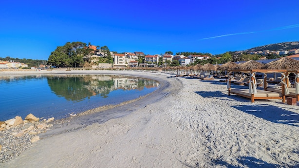 A beach with a clear blue sky, a sandy shore, and a row of thatched-roof structures with seating. - House Nonina Kuca (Rab)