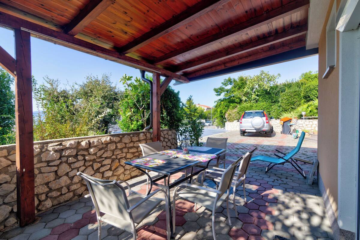 Eine Terrasse im Freien mit einer Steinmauer, einer Holzhütte, einem Tisch mit Stühlen und einem Parkplatz im Hintergrund.