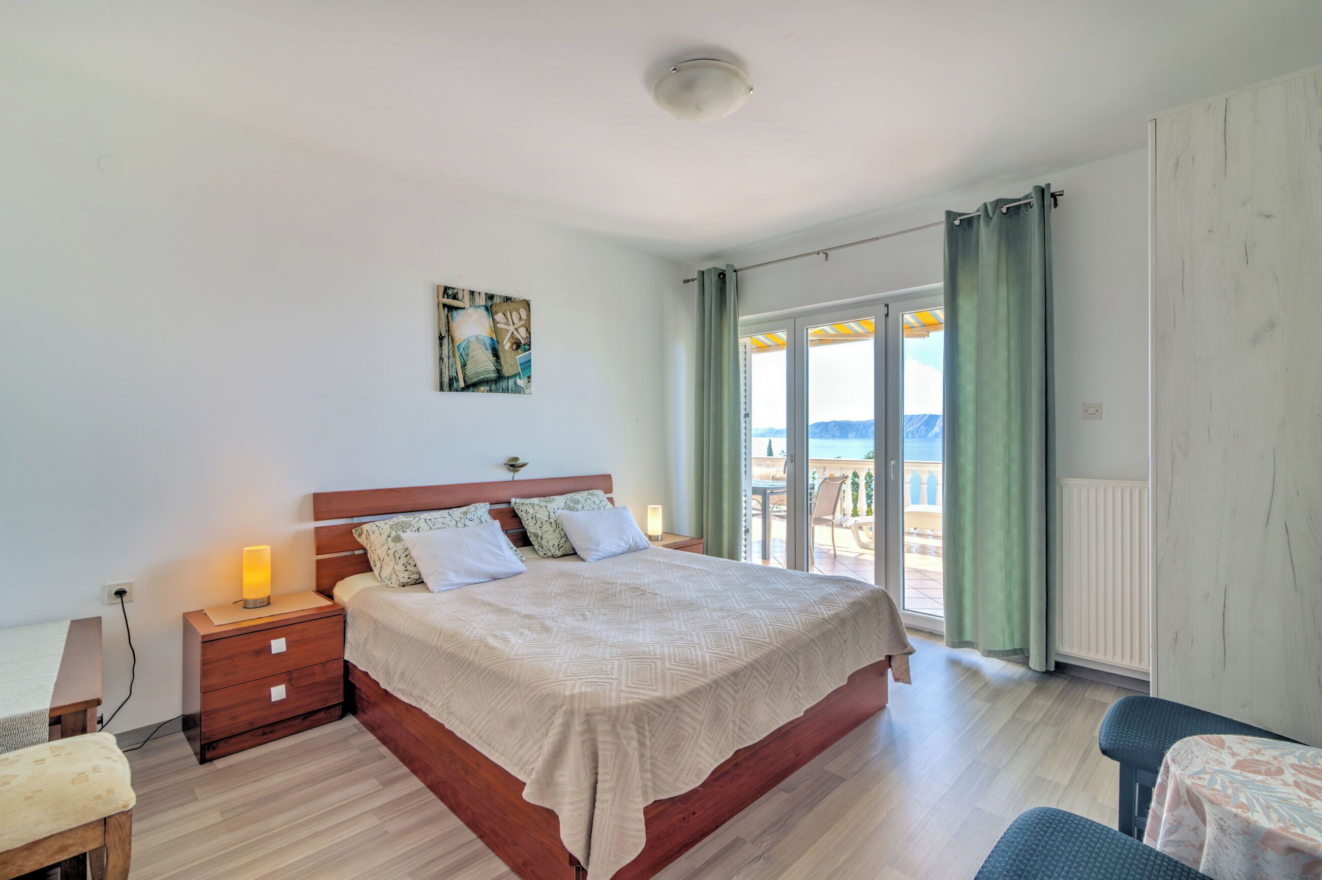 Un dormitorio con una cama de madera, una mesita de noche con lámpara, una pequeña mesa y vistas al exterior a través de un ventanal corredero.