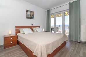 A bedroom with a large bed, a nightstand, a painting, and a view of the outdoors through a sliding glass door. - Apartments Dino & Toni (Novi Vinodolski)