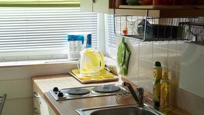 Een keuken met een spoelbak, fornuis en kastjes. Er is een raam met jaloezieën, een afwasschaal en keukengerei.
