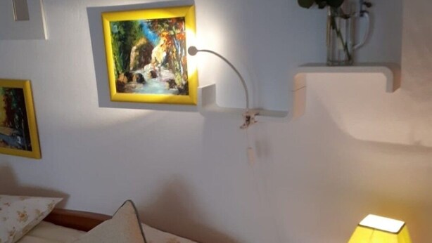 Een slaapkamer met een bed, een bedlampje, een lijst met een afbeelding aan de muur en een kleine plant op een plank.