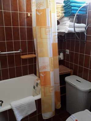 A bathroom with a bathtub, a shower curtain, a towel rack with folded towels, a waste basket, and a small wooden stool. - Apartment Apartment Yellow (Baska Voda)