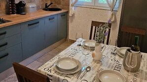 Un comedor con una mesa preparada para cuatro personas, una cocina con armarios de madera y un ventanal con cortinas translúcidas.
