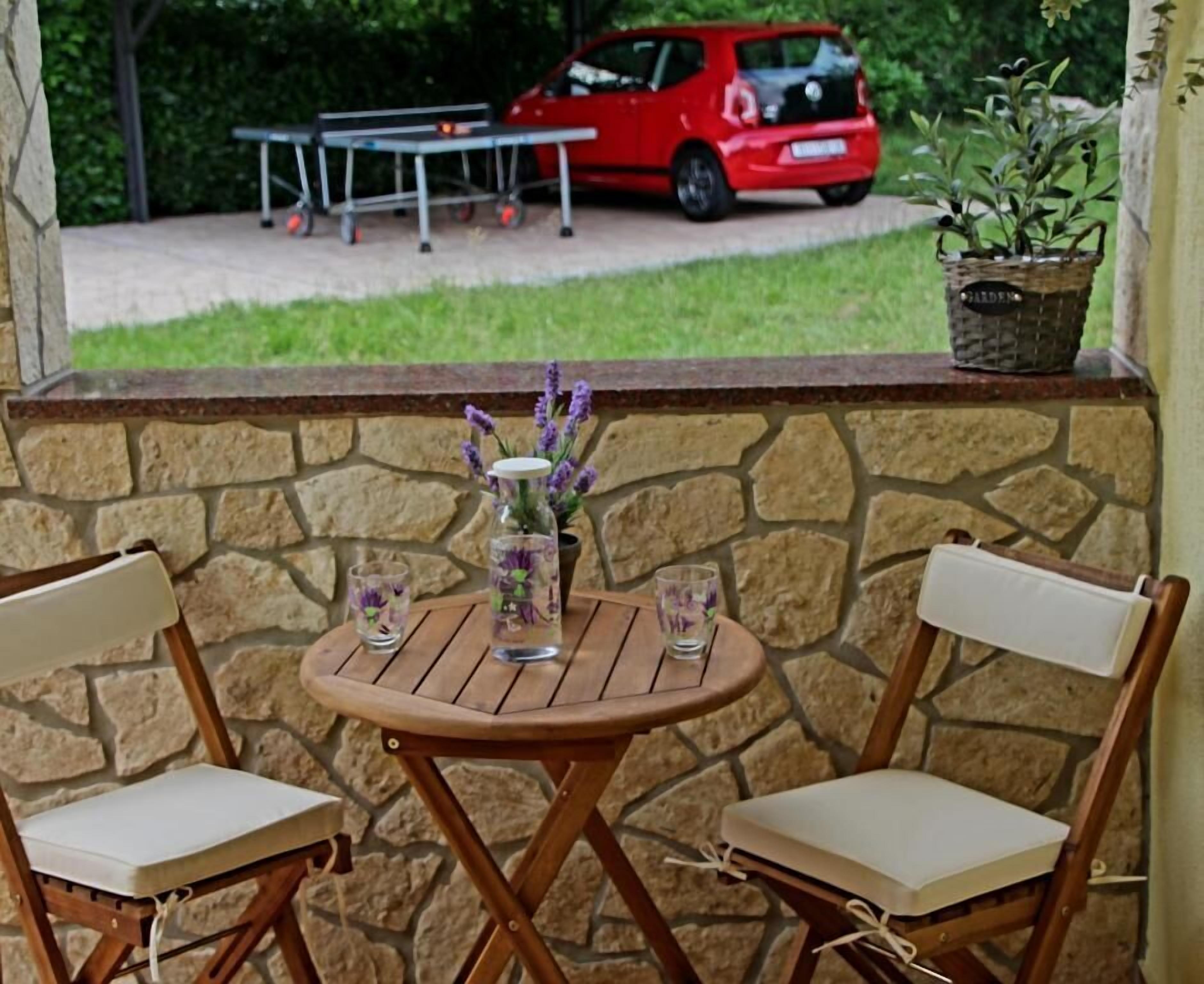 Um pátio com parede de pedra, duas cadeiras de madeira, uma mesa redonda pequena e um vaso com flores.