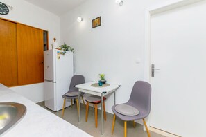 A compact kitchen with a white refrigerator, a wooden door, and a small dining table with two chairs. - Apartment New (Supetar)