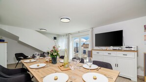 A dining area with a long wooden table set for six, white plates, wine glasses, and a bottle of white wine. There are black chairs around the table, and a flat-screen TV mounted on the wall. - House Emma (Crikvenica)