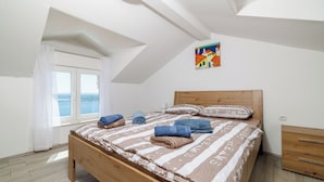 A bedroom with a wooden bed, a window with white curtains, a painting on the wall, and a wooden nightstand. - House Emma (Crikvenica)