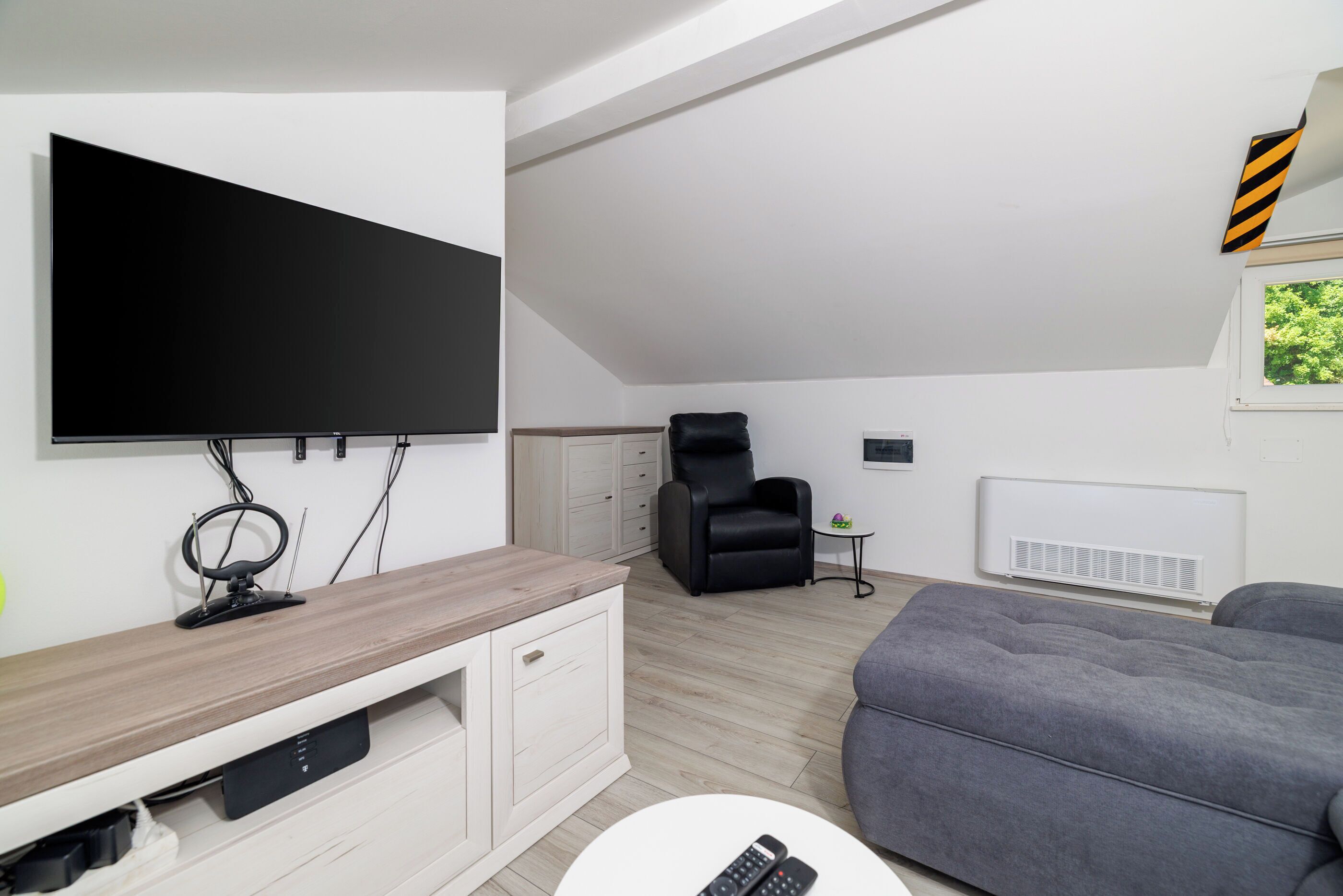 A modern living room with a flat-screen TV mounted on the wall, a grey sofa, a wooden TV stand, and a black chair.