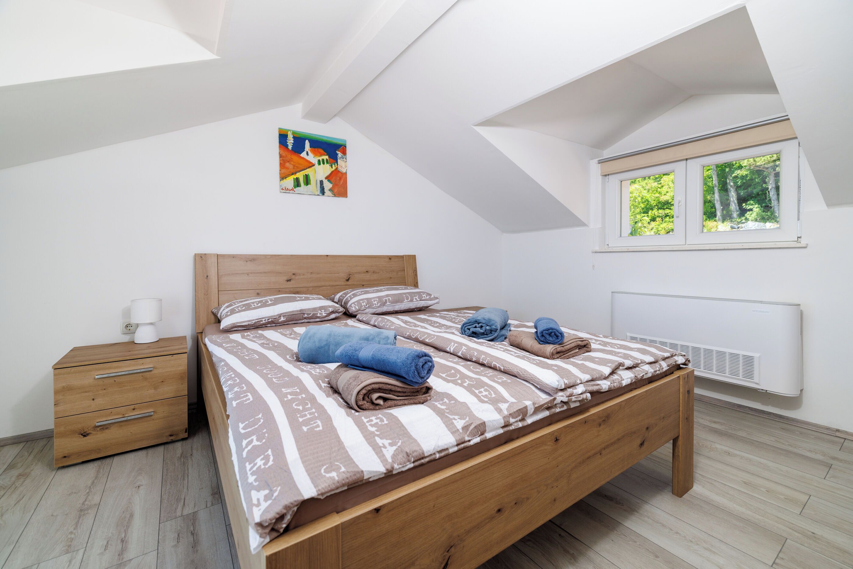A bedroom with a wooden bed, a nightstand, a radiator, and a window with a view of greenery.