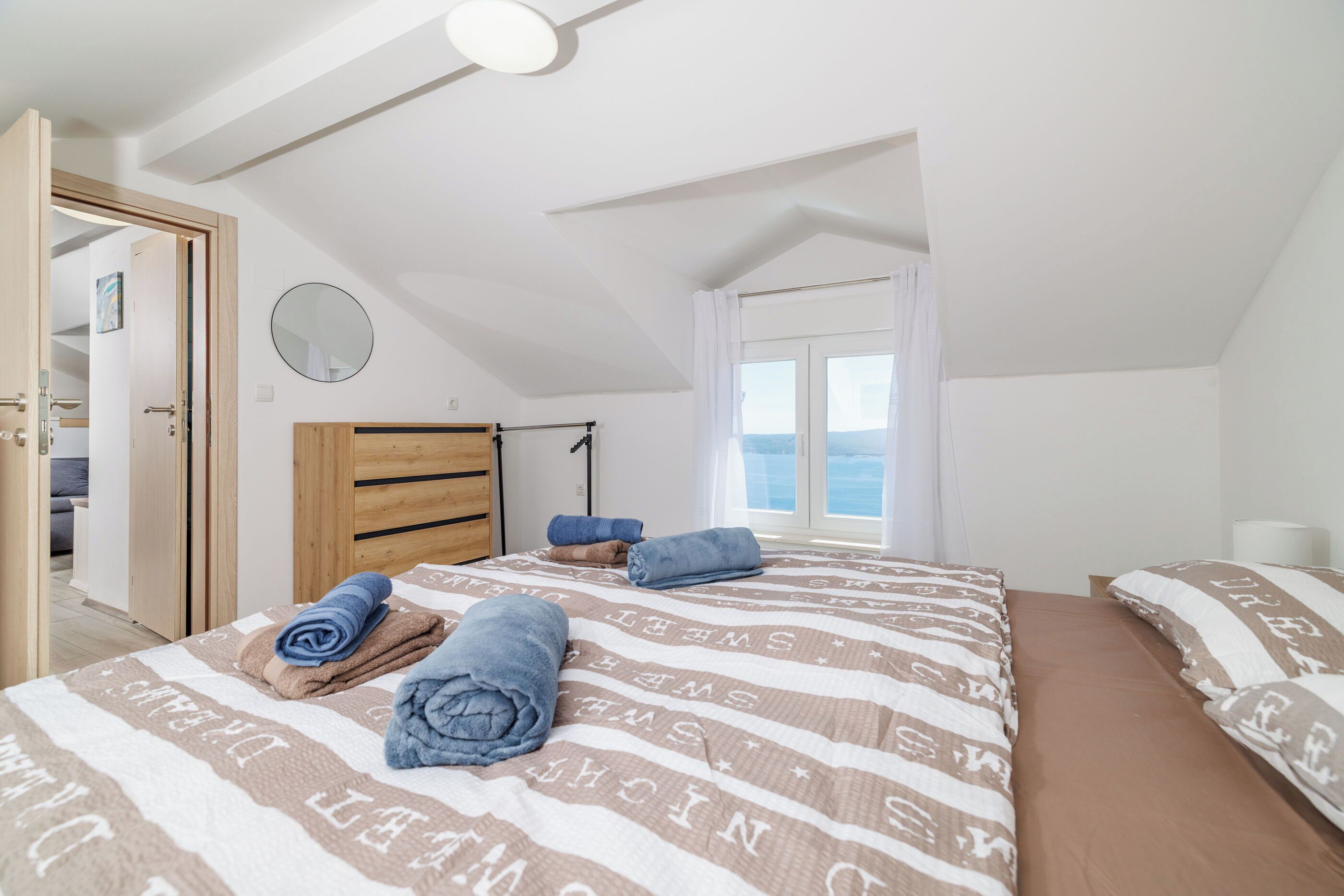 A bedroom with a sloped ceiling, a wooden wardrobe, a bed with striped bedding, and a window with curtains.