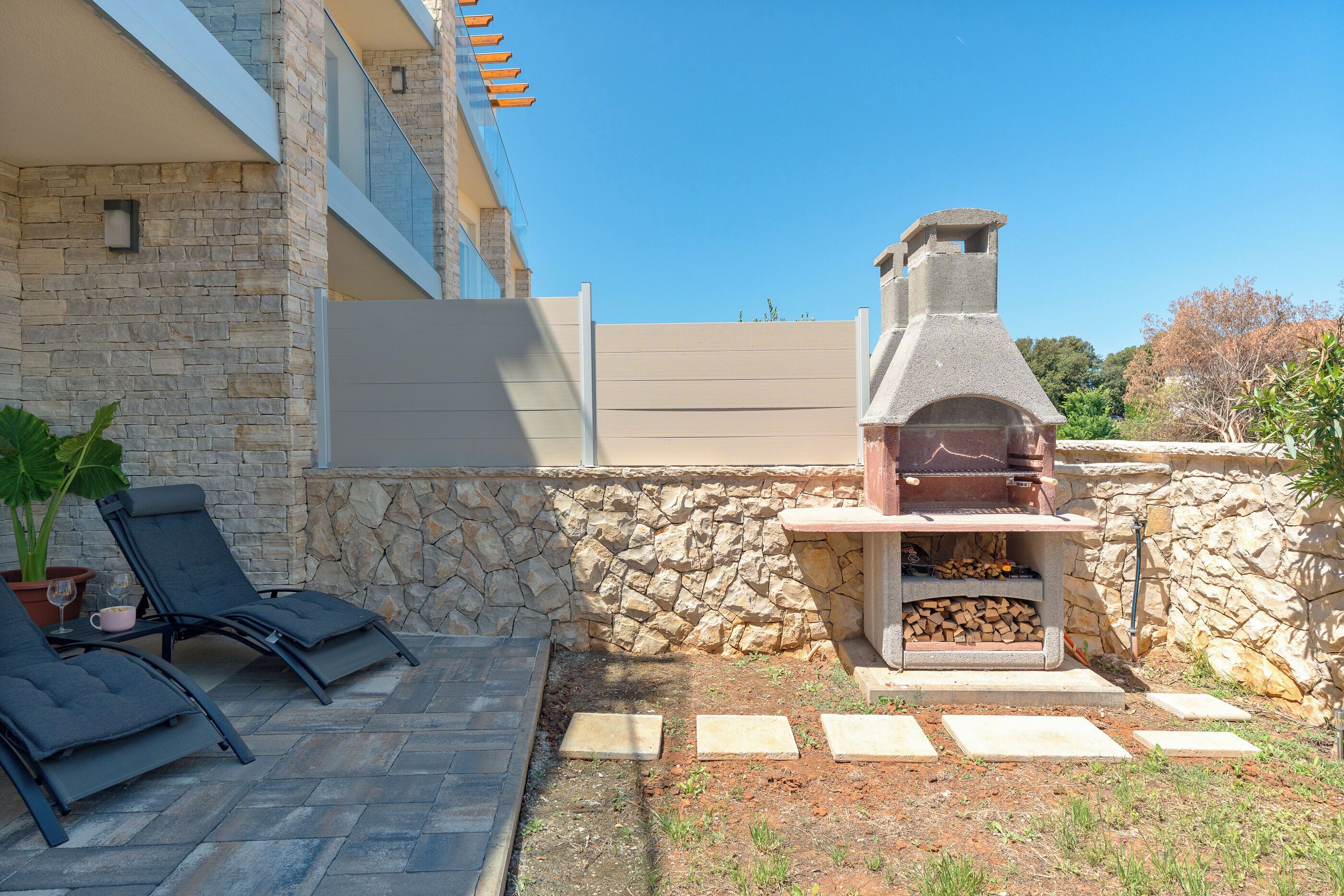 A stone wall with a barbecue grill, lounge chairs, and a potted plant.