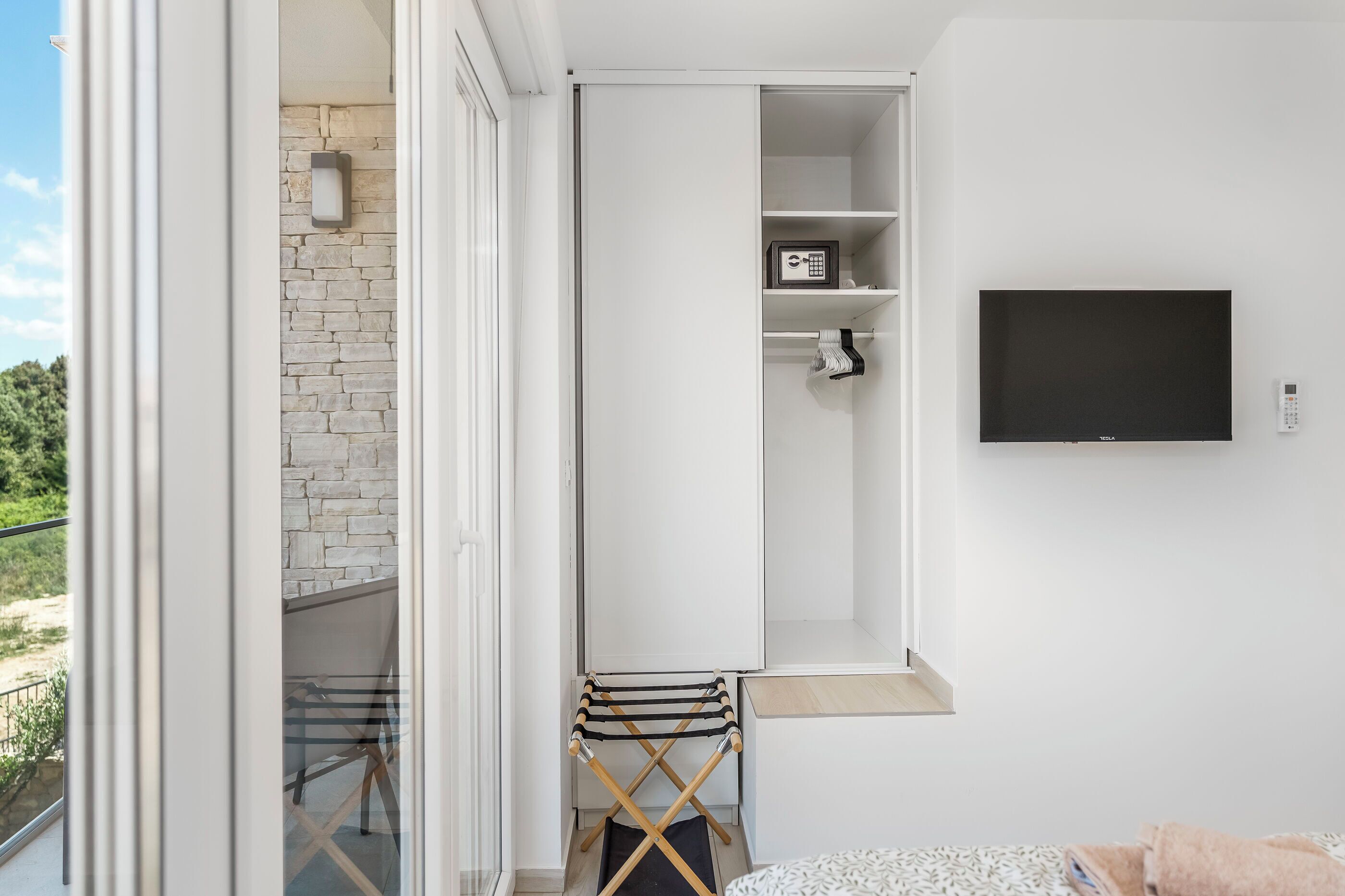 A modern, compact room with a sliding door leading to a balcony, a mounted flat-screen TV, a small seating area, and a built-in safe.