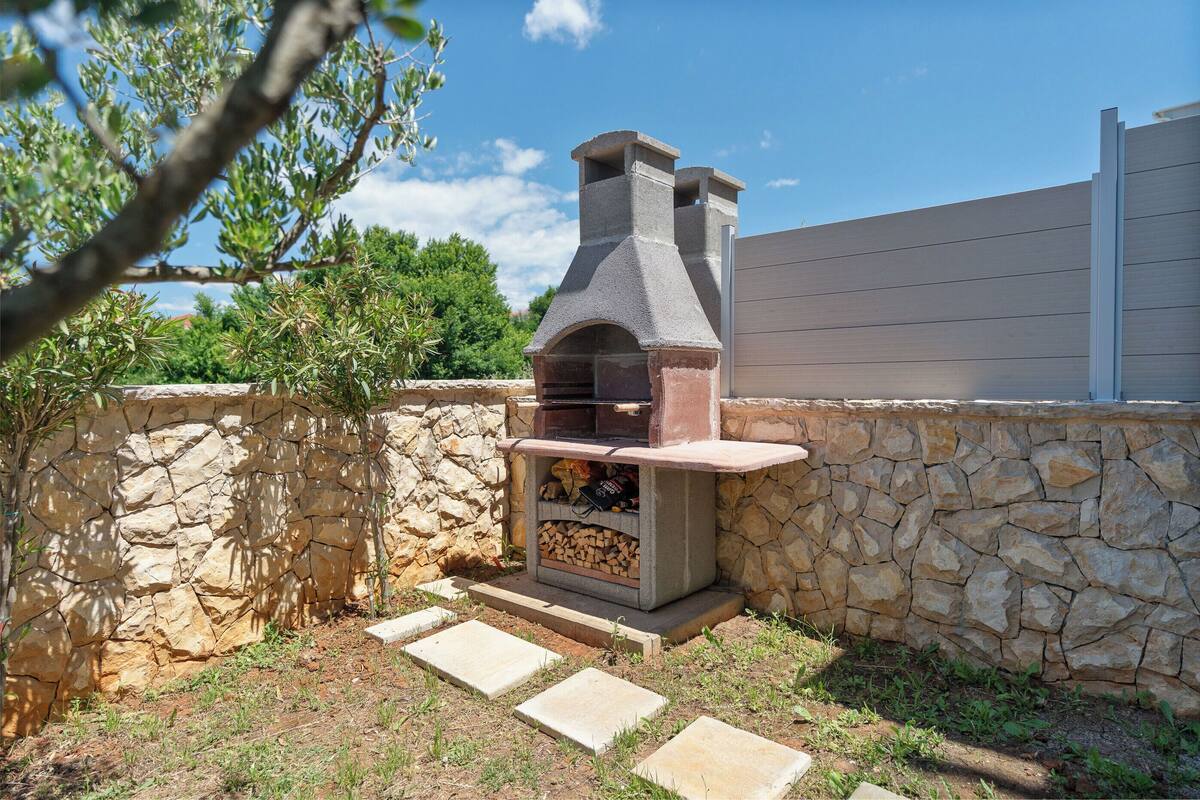 An outdoor stone barbecue grill with a chimney, set against a stone wall and a wooden fence.