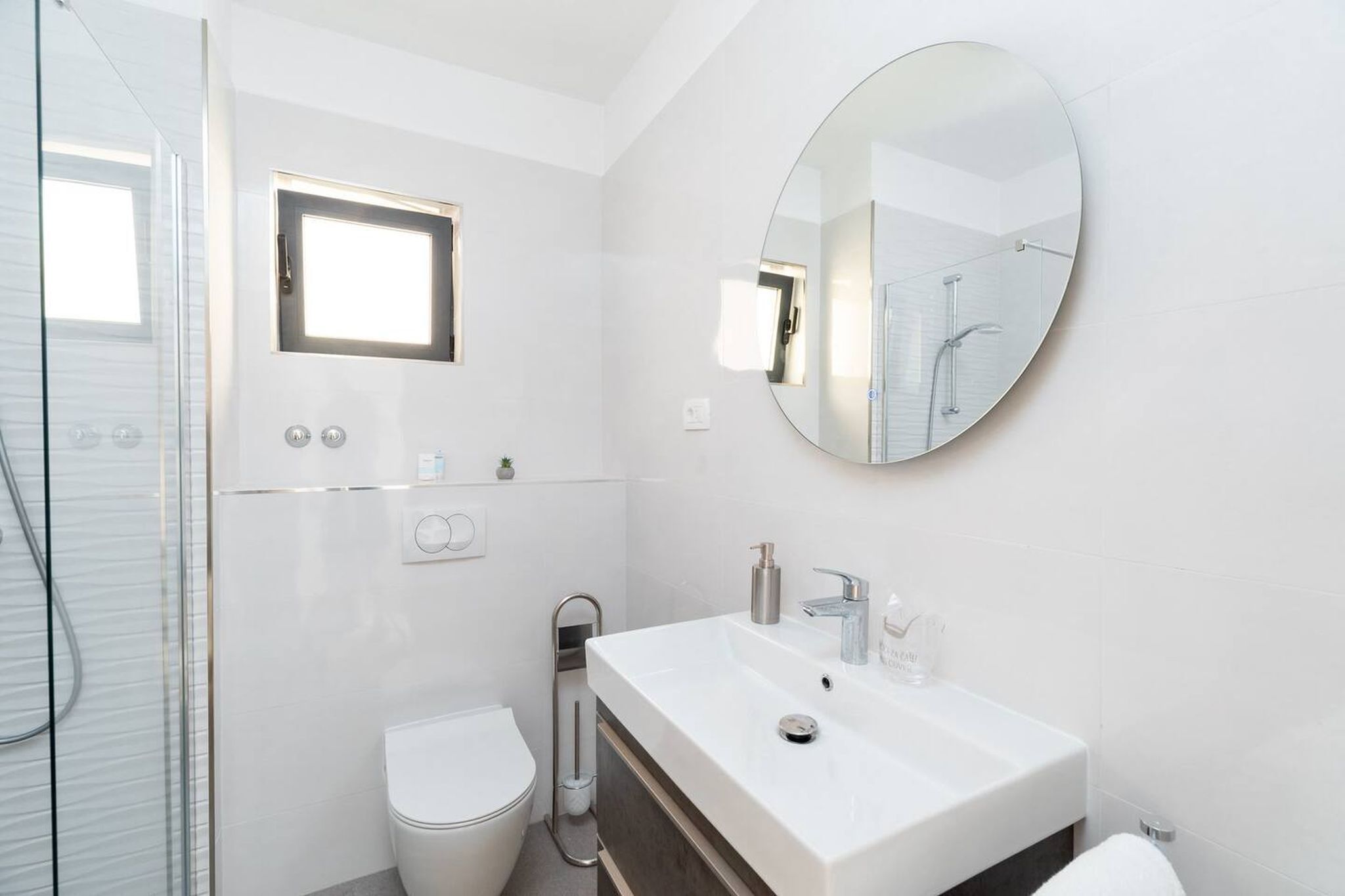 A modern bathroom with a glass shower enclosure, a white sink with a round mirror, and a toilet.