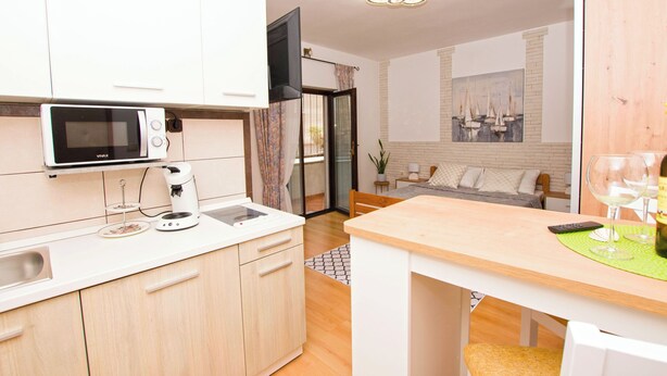 Een moderne keuken met witte kasten, een magnetron en een koffiezetapparaat. Er is een eethoek met een tafel en stoelen, en een woonkamer met een bank en een schilderij aan de muur.