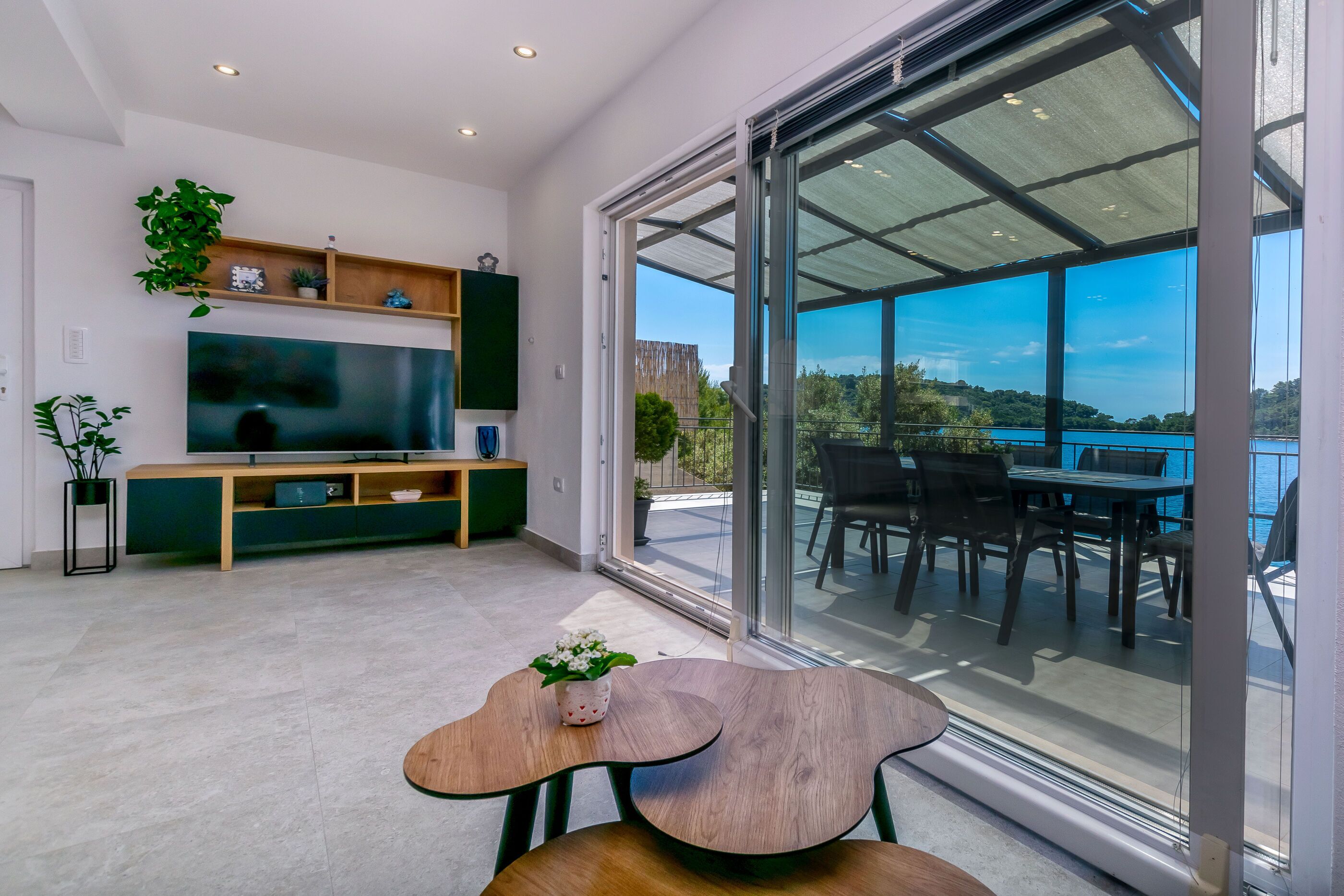 A modern living room with a flat-screen TV, wooden furniture, and a balcony with a table and chairs overlooking a body of water.