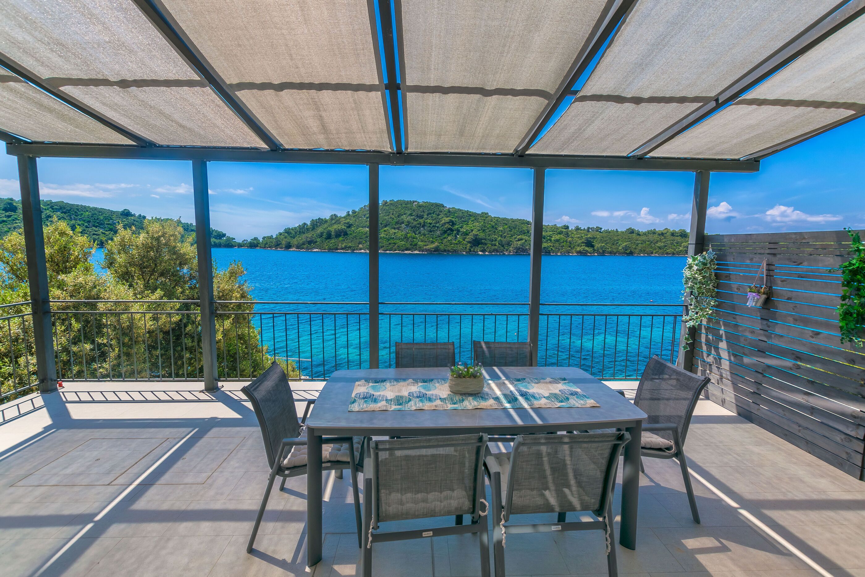 Uma área de estar coberta ao ar livre, com uma mesa e cadeiras, com vista para um corpo de água e uma encosta verde.