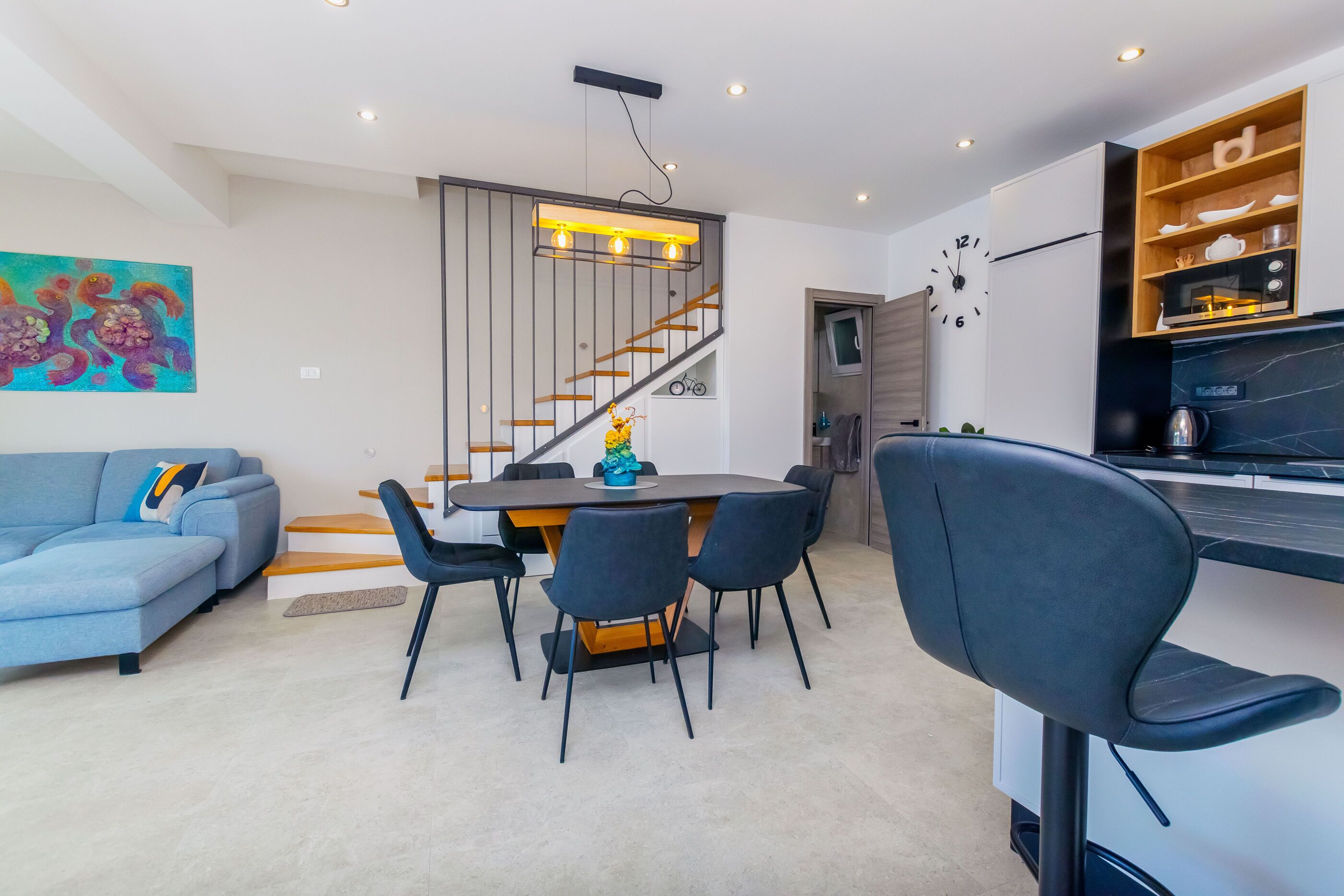 A modern living room with a sofa, dining table, chairs, a staircase, and a kitchen area with a microwave.