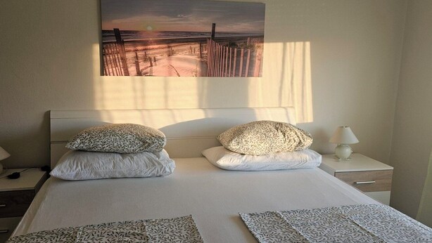 A hotel room with a large bed, two bedside tables, a lamp, and a beach scene painting on the wall. - Apartments Madeji (Rab)