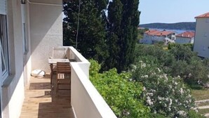 A balcony with a tiled floor, a small table, and a chair. There are potted plants and a view of a coastal area with buildings and trees. - Apartments Madeji (Rab)