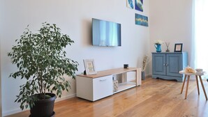 Sala de estar con piso de madera, mueble blanco para equipo multimedia, televisor de pantalla plana grande, gabinete azul y planta en maceta.
