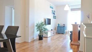 Un salón espacioso con piso de madera, una mesa de comedor con sillas, una planta en maceta, un mueble azul y un televisor en la pared.