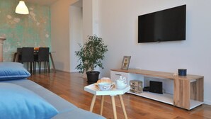 Una sala moderna con un sofá, una mesa de comedor con sillas, una planta en maceta, un televisor en la pared y un mueble bajo de madera con una bandeja de objetos.