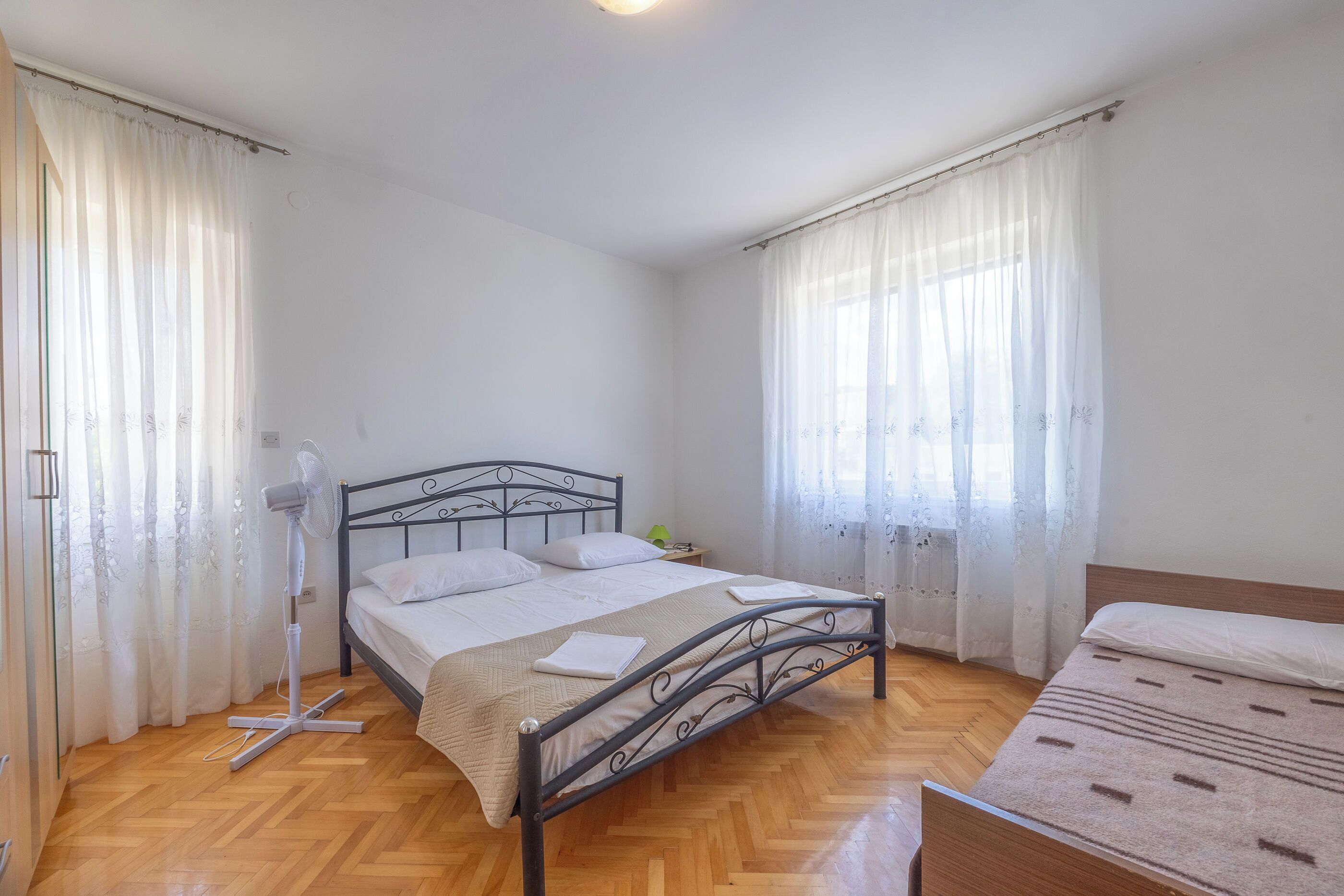 Une chambre à coucher avec un lit, un ventilateur sur pied et une fenêtre avec des rideaux.