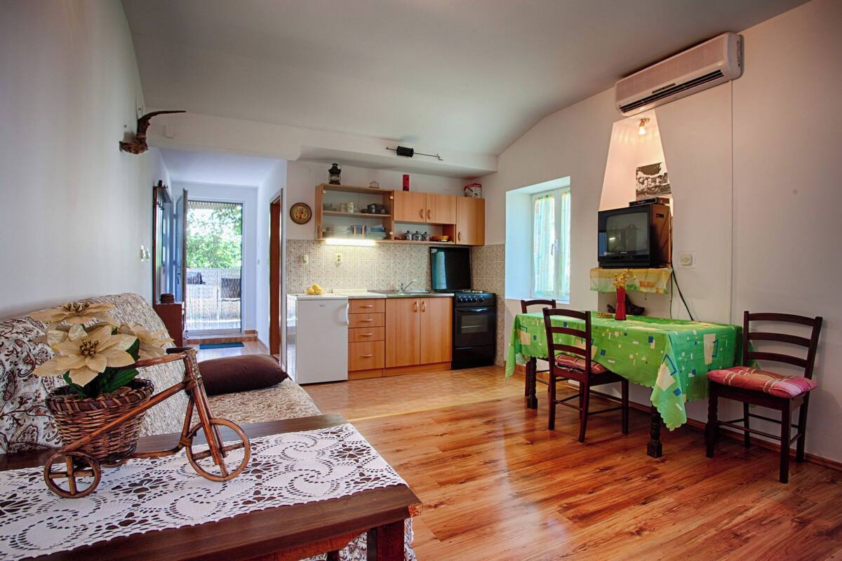 A compact kitchen with wooden cabinets, a refrigerator, and a stove. There is a dining table with chairs and a television on a stand.