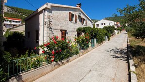A stone building with a sloped roof, a small porch, and a garden with red flowers. - Apartments Dina (Komiza)
