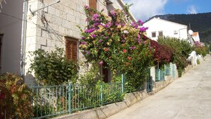 Un edificio de piedra con puerta de madera y ventana, rodeado por una cerca de metal y plantas con flores.