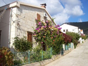 A stone building with a wooden door and a window, surrounded by a metal fence and flowering plants. - Apartments Dina (Komiza)