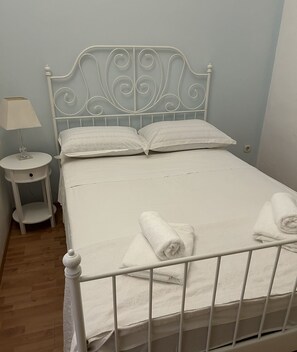 En pænt redt seng med hvide sengetøj og puder, et lille natbord med en lampe, og et foldet håndklæde på sengen.