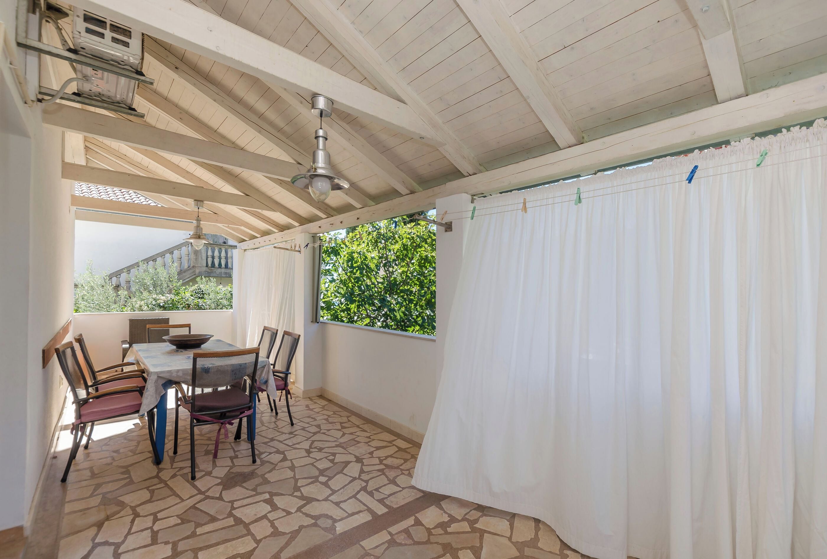 A covered patio area with a table and chairs, a ceiling fan, and a view of greenery outside.