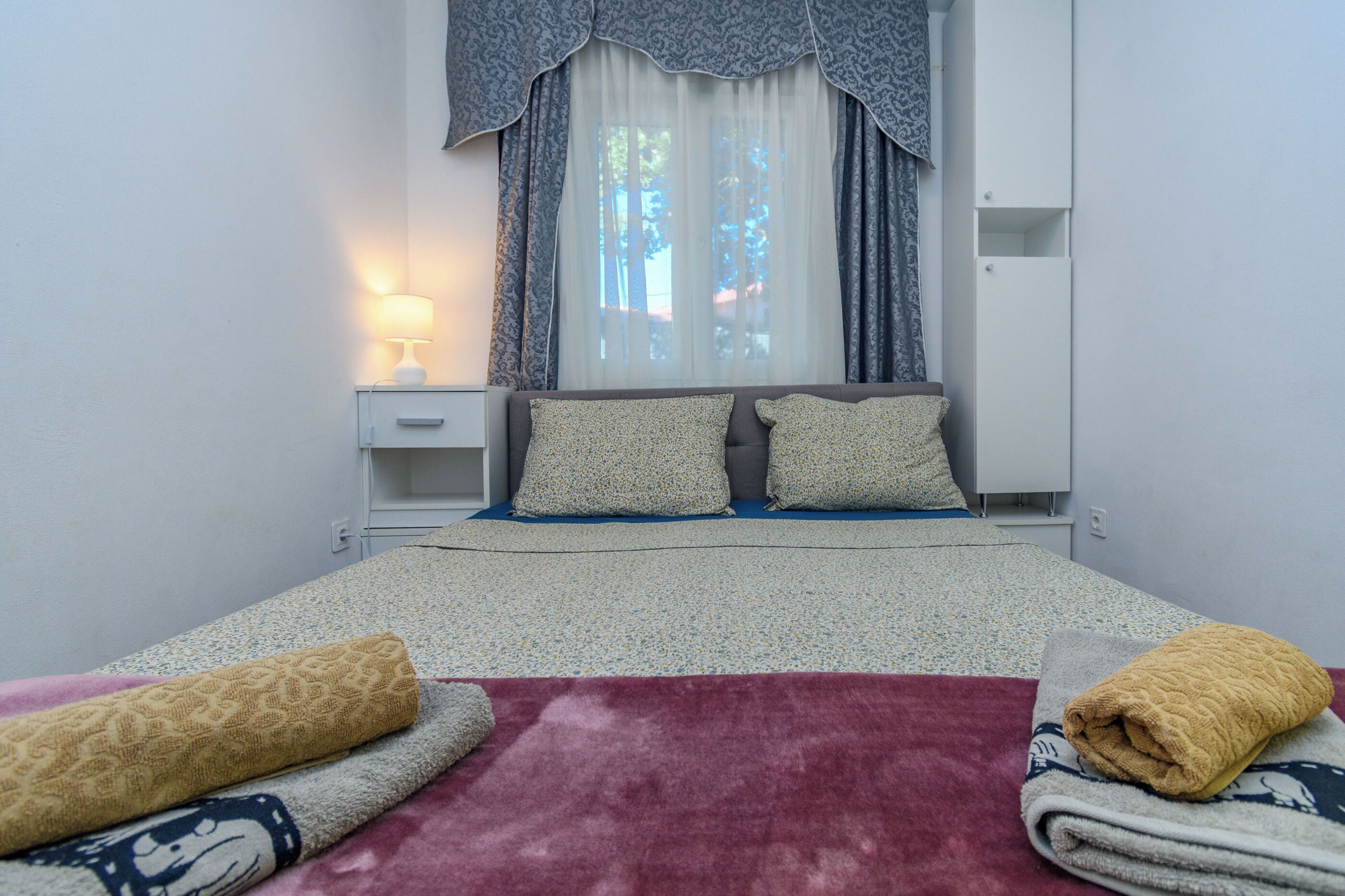 A bedroom with a bed, a nightstand, a lamp, a wardrobe, and a window with curtains.