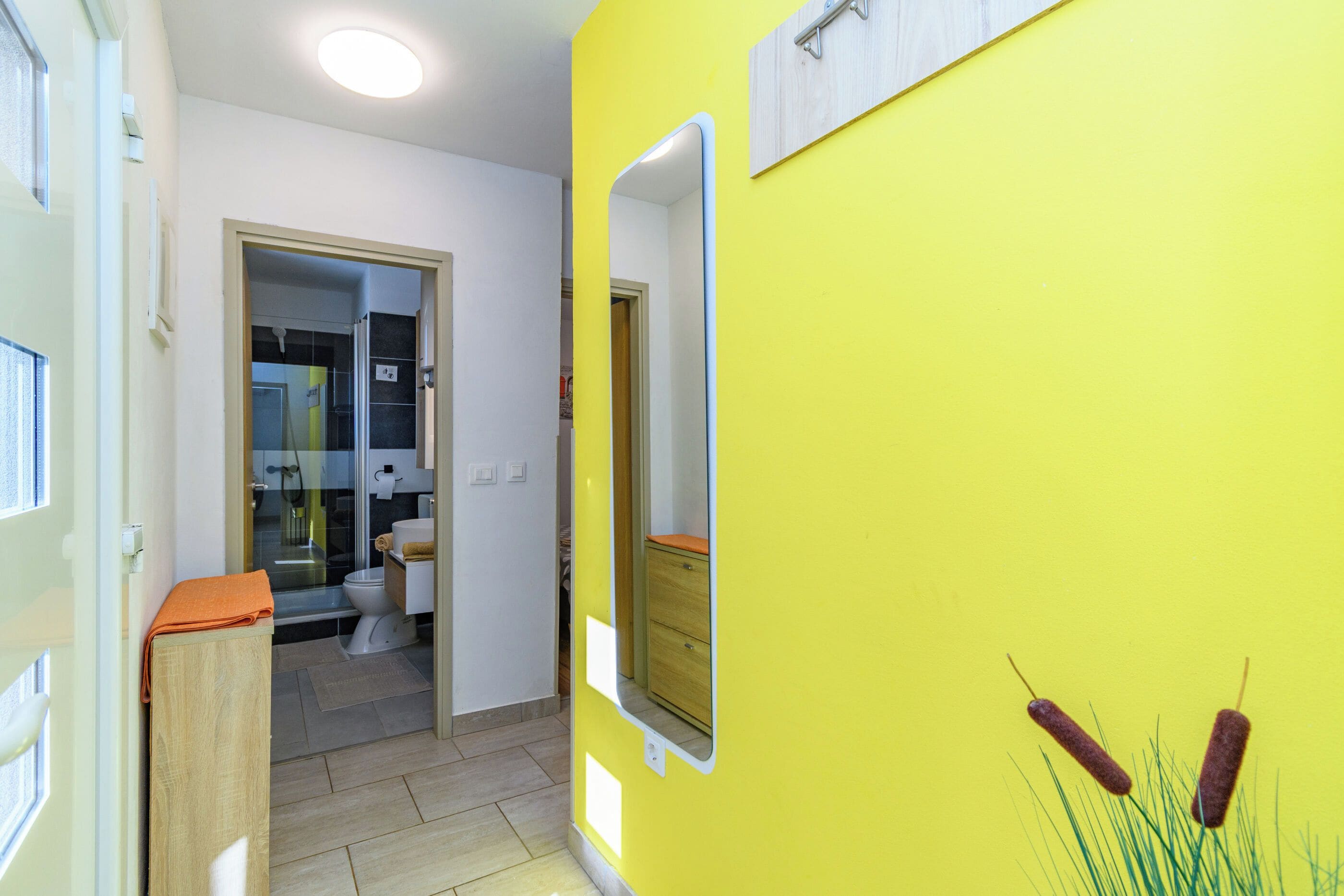 A hallway with a yellow wall, a mirror, and a door leading to a bathroom with a toilet and shower.