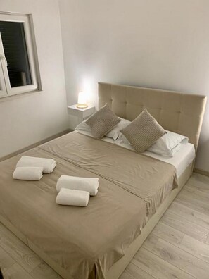 A neatly made bed with a beige bedspread and pillows, a bedside lamp, and rolled towels on the floor. - Apartment Julia (Tisno)