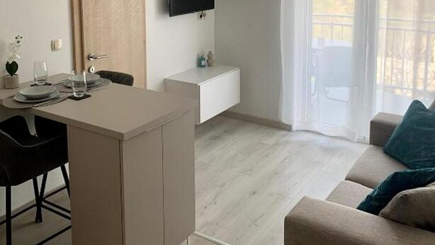 A modern living room with a sofa, a dining area with a table and chairs, a TV mounted on the wall, and a balcony with curtains. - Apartment Julia (Tisno)