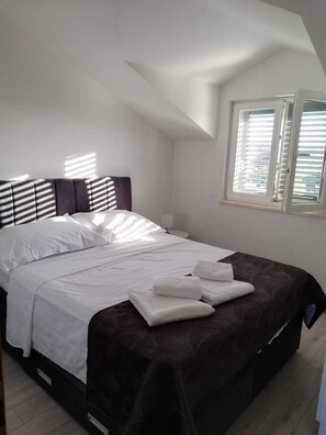 Quarto com cama, roupa de cama branca, colcha marrom escura e janela com persianas.
