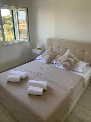 A neatly made bed with white linens and pillows, a bedside table with a lamp, and a window with a view of greenery. - Apartment Julia (Tisno)