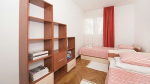 Een slaapkamer met een houten boekenkast, twee bedden met beddengoed met een patroon, een raam met dunne gordijnen en een houten vloer.