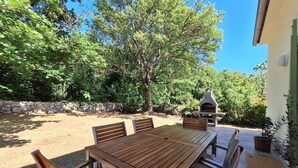 En udendørs terrasse med et træbord, stole, en stenvæg og en grill.