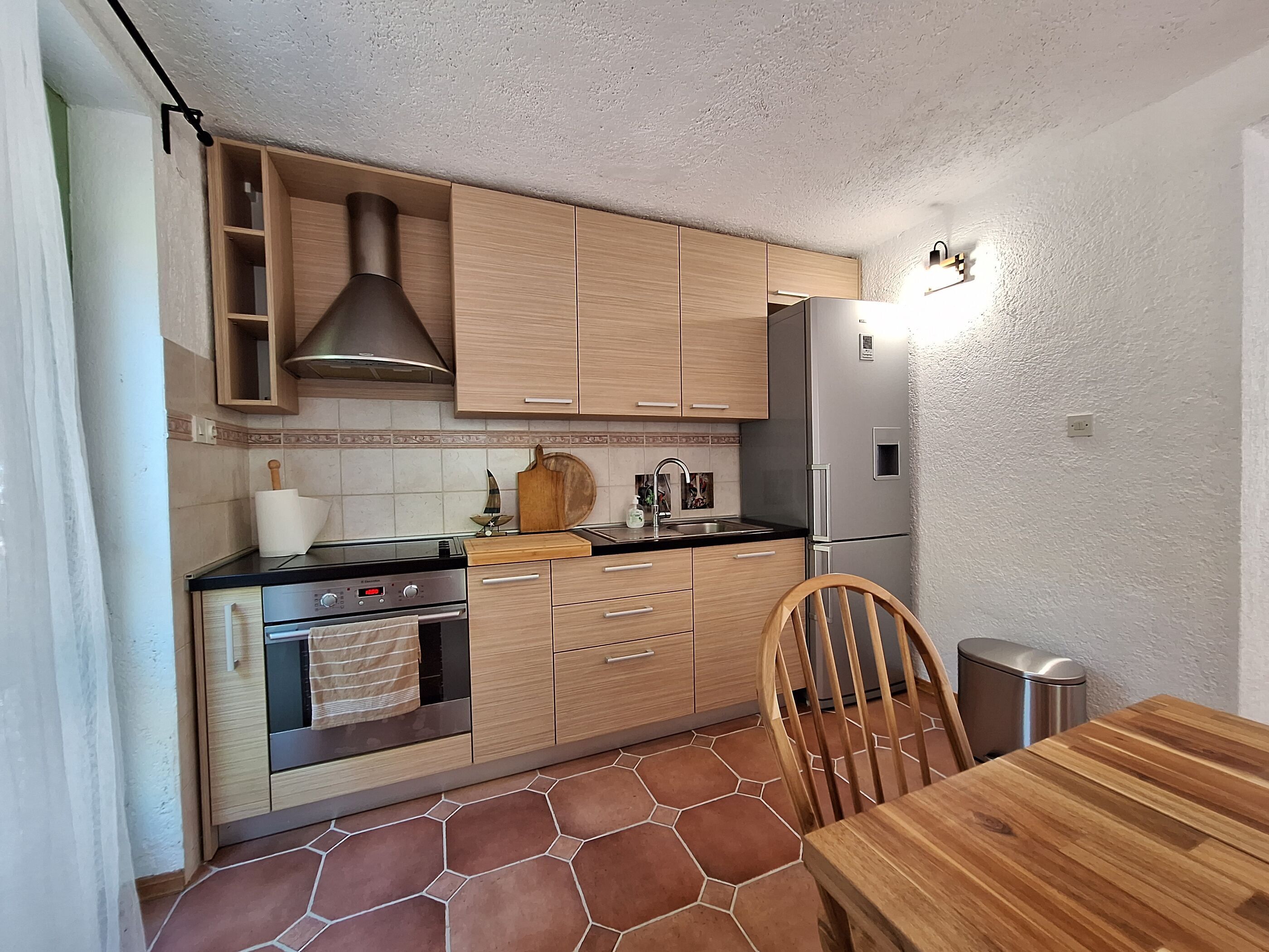 Une cuisine compacte dotée de meubles en bois, d’un réfrigérateur en acier inoxydable, d’une cuisinière à gaz et d’une table à manger avec des chaises.