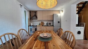 Et træbord med stole, en dekorativ skål i midten, en hængende lampe og et køkken med skabe og hårde hvidevarer.