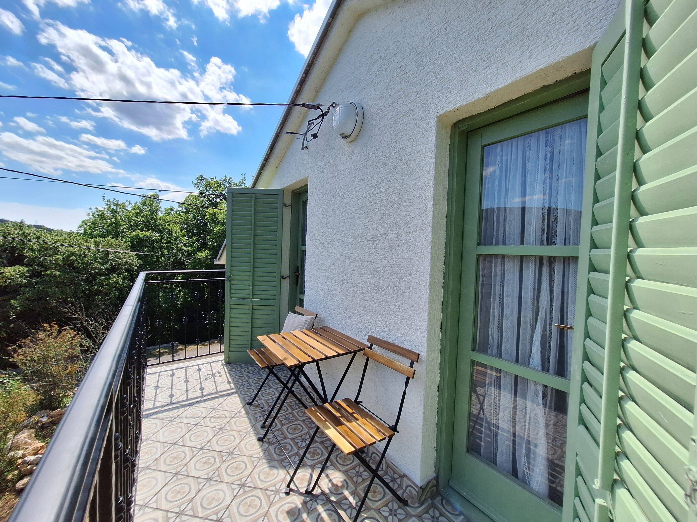 A balcony with two wooden chairs and a table, a window with green shutters, and a patterned tile floor.